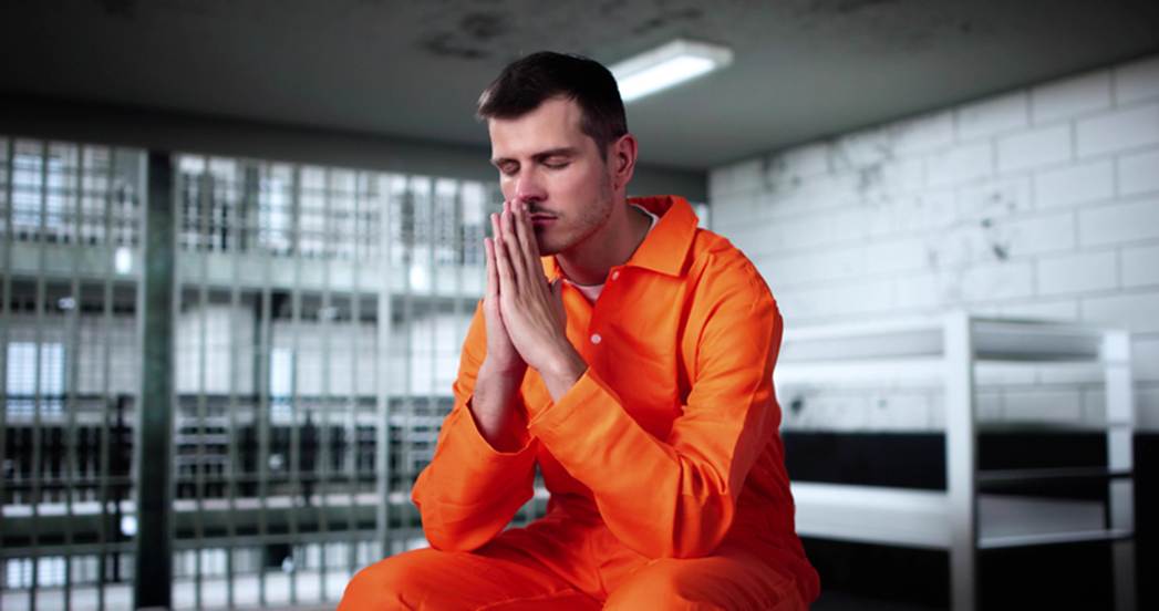 A man in an orange jumpsuit, eyes closed and hands pressed together, sits in a jail cell. This image illustrates the consequences of false imprisonment.